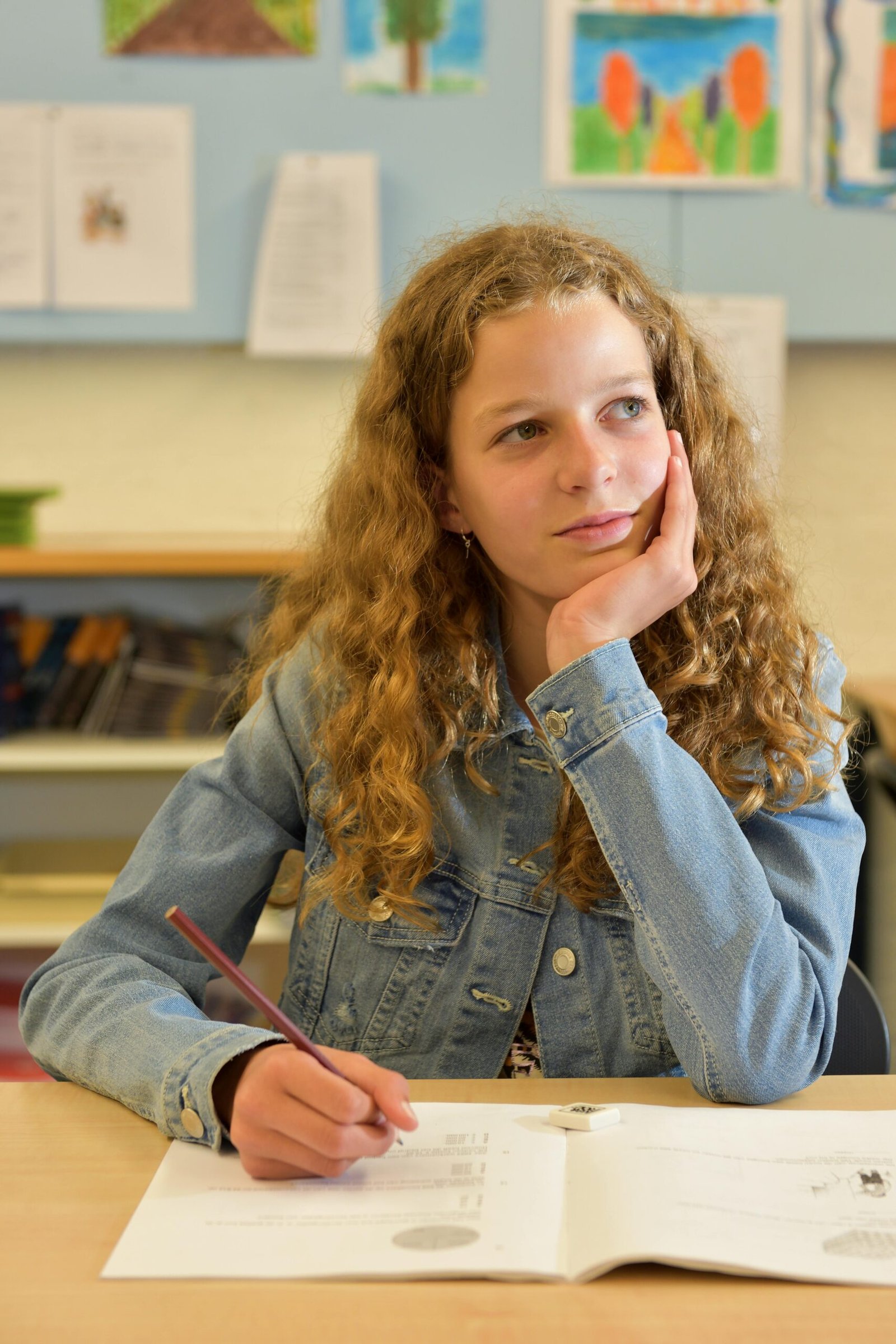 Thoughtful teenage girl in a classroom, holding a pencil and writing in a notebook.