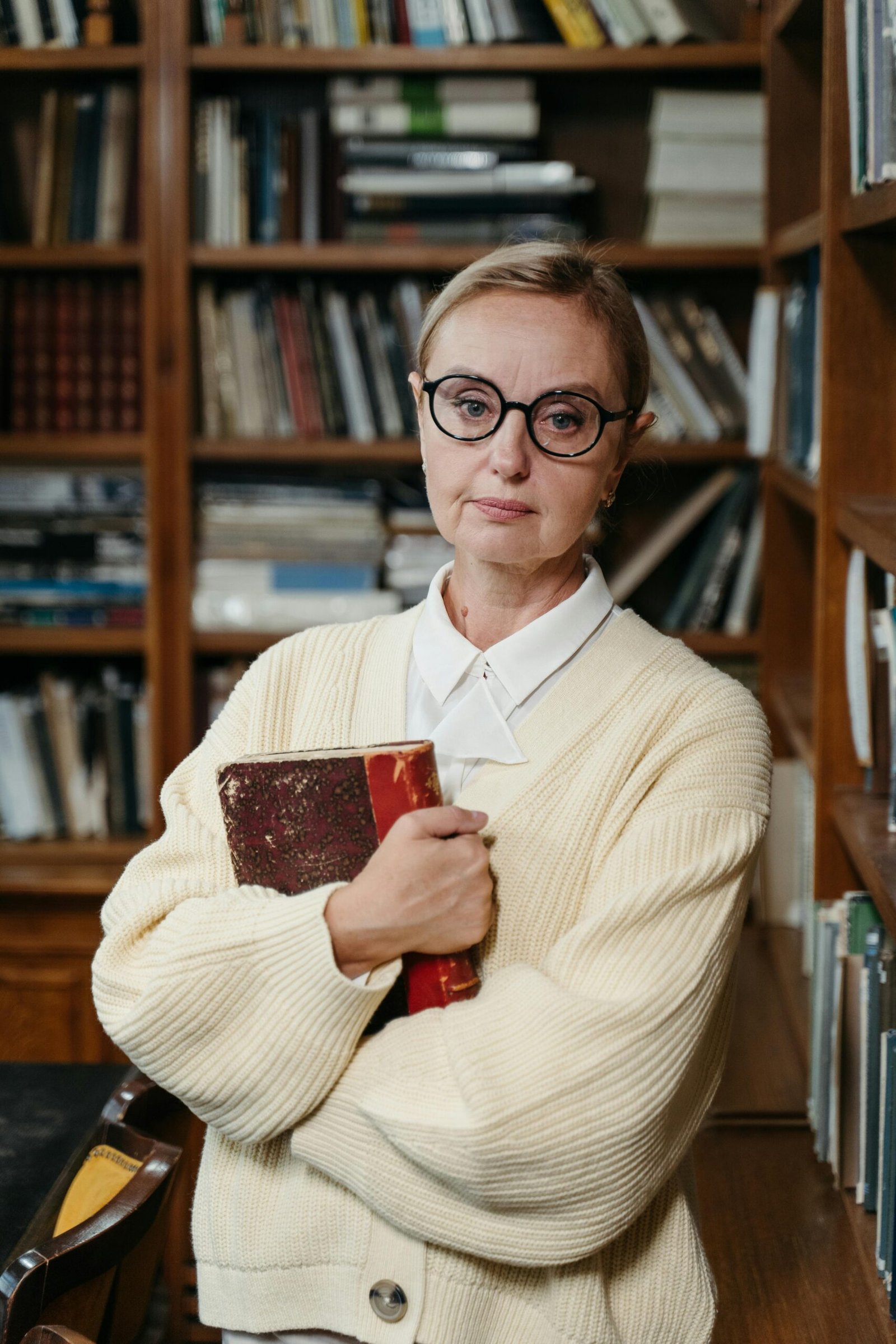 Confident woman in eyeglasses holding a book in a library setting, showcasing style and knowledge.